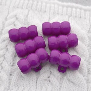 May include: Five purple plastic hair clips, each with three rounded sections, are arranged on a white cable-knit fabric. The clips have a glossy finish and a small loop at the base.
