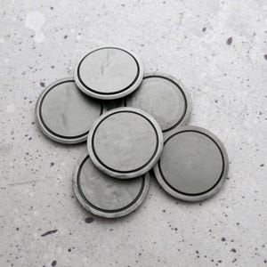 May include: A collection of seven silver-colored, round metal objects with a black border. The objects are arranged in a slightly overlapping pattern against a speckled gray background. The metal has a matte finish.