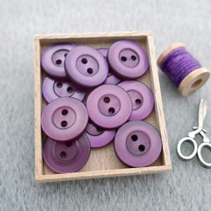 May include: A collection of round, two-hole purple buttons contained within a small, square wooden tray. Beside the tray sits a wooden spool wrapped with purple thread and a pair of small, silver-colored metal scissors with ring handles.