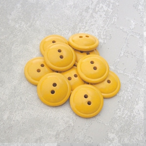 May include: A collection of yellow, round buttons with two holes each. The buttons are clustered together, showcasing their uniform shape and color. The buttons are made of a smooth material.