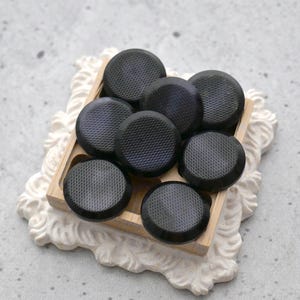 May include: A collection of eight black, round buttons with a textured surface, arranged in a wooden tray. The tray sits on a decorative, white, ornate base. The buttons have a slightly domed shape.