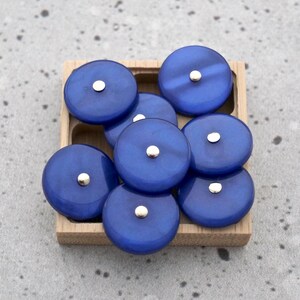May include: A collection of deep blue, circular beads with a metallic silver center, arranged in a small, light-colored wooden tray. The beads have a glossy finish and are evenly spaced within the tray.