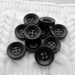 May include: A collection of dark gray, round buttons with four holes each. The buttons are arranged on a white, textured fabric, likely a knitted or crocheted material. The buttons have a slightly concave design.
