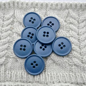 May include: A collection of eight round, matte navy blue buttons, each featuring four central holes for attachment. The buttons are arranged in a cluster, resting against a light gray knitted fabric with a visible vertical cable stitch pattern.