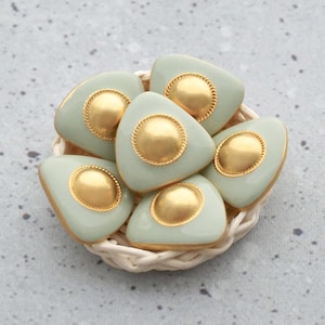 May include: A collection of six light green, triangular-shaped decorative objects with gold accents, arranged in a small woven basket. Each piece features a gold center and gold trim, creating a sophisticated aesthetic.