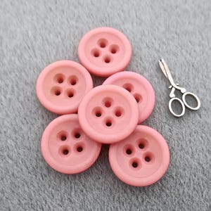 Puede incluir: Seis botones redondos de color rosa claro con cuatro agujeros cada uno, junto con unas pequeñas tijeras plateadas. Los botones están agrupados sobre una superficie gris, ideales para proyectos de costura y manualidades.