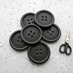 May include: A collection of seven black buttons with four holes each, arranged in a pile. A small pair of silver and black scissors rests nearby. The buttons have a matte finish and are on a light gray surface.