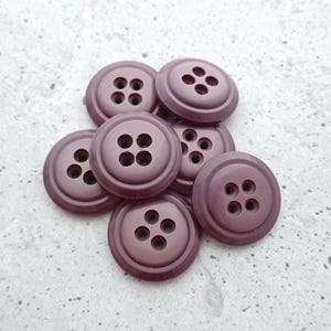 May include: A collection of seven round, dark mauve buttons, each featuring a smooth, slightly raised center and a recessed edge. Every button has four evenly spaced holes in the middle for attachment, arranged in a square pattern on the surface.