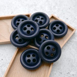 May include: A collection of navy blue buttons with four holes each, arranged in a wooden tray. The buttons have a classic, round shape and a matte finish. The buttons are a versatile sewing supply.