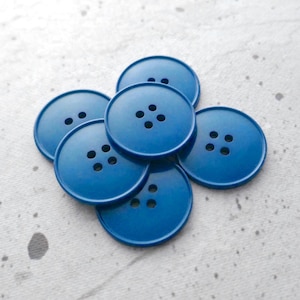 May include: A collection of six round, solid blue buttons arranged in a cluster. Each button features a smooth, flat surface with four small, evenly spaced holes in the center for attachment. The buttons have a consistent, deep blue color.