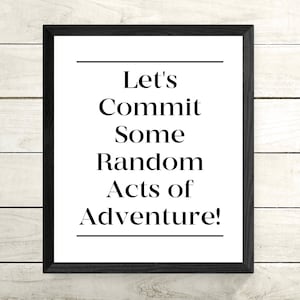 May include: A framed print with the text "Let's Commit Some Random Acts of Adventure!" in a black font. The print is in a black frame and is set against a white wooden background.