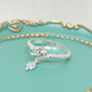 May include: A silver snake ring with a dangling charm. The snake is coiled and has a pink stone for an eye. The charm is a leaf with a clear stone.