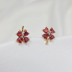 Może przedstawiać: Para złotych kolczyków z czerwonym wzorem koniczyny czteroliściennej. Każda koniczyna ma pięć czerwonych kamieni w kształcie serca i jeden przezroczysty kamień w środku.