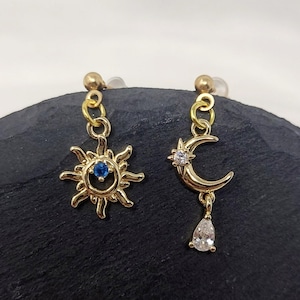 May include: Gold-toned mismatched earrings featuring celestial designs. One earring showcases a sunburst with a blue center stone, while the other features a crescent moon with a star and a dangling teardrop-shaped gem. The earrings are displayed on a dark surface.