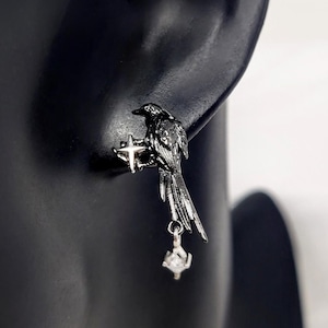 May include: A pair of black raven earrings. Each earring features a detailed raven perched on a cross-shaped design, with a dangling, clear, cubic zirconia stone. The earrings are made of a dark metal and are displayed on a black mannequin.