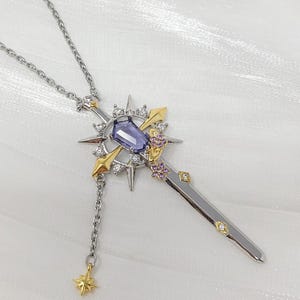 May include: A silver pendant necklace with a sword-shaped design. The sword features a purple gemstone, gold details, and small pink and clear gems. A chain with a star charm hangs from the sword.