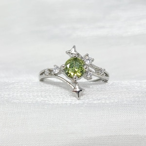May include: A silver ring featuring a central, round, light green gemstone, surrounded by smaller clear stones and star-shaped accents. The ring has a delicate, celestial design, set against a white background.