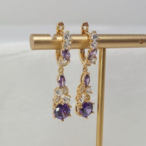 Può includere: Un paio di orecchini a cerchio color oro con pietre preziose viola e bianche pendenti. Gli orecchini presentano una grande pietra viola rotonda nella parte inferiore, con pietre viola e bianche più piccole che salgono verso il cerchio. Gli orecchini sono esposti su un supporto dorato.