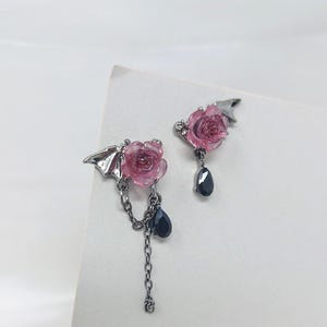 May include: A pair of gothic-style earrings featuring pink rose-shaped pendants with black teardrop accents. The earrings have silver-colored bat wing details and chain attachments, creating a unique and elegant design.
