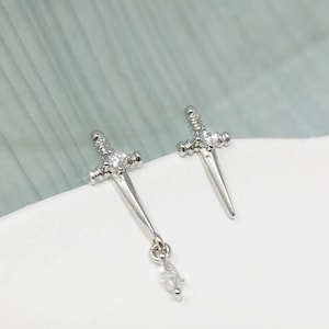 May include: A pair of silver-coloured earrings shaped like miniature swords. One is a stud, the other has a dangling, teardrop-shaped charm. Both earrings feature small, clear gemstones.