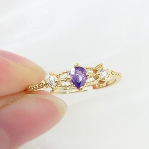May include: A gold-colored ring featuring a pear-shaped purple gemstone at the center, flanked by two smaller white gemstones. The ring has a delicate, openwork design with a textured band.
