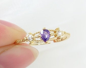 May include: A gold-colored ring featuring a pear-shaped purple gemstone at the center, flanked by two smaller white gemstones. The ring has a delicate, openwork design with a textured band.