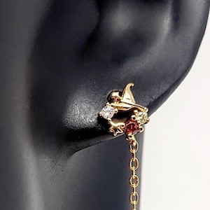 Peut inclure: Boucles d'oreilles pendantes dorées avec un groupe de pierres précieuses rouges, blanches et transparentes. Les boucles d'oreilles ont une chaîne délicate qui pend du groupe.