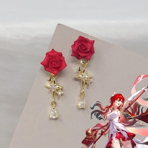 May include: A pair of gold-toned earrings featuring a red rose stud at the top, followed by a dangling gold star charm accented with clear crystals and a small, teardrop-shaped clear crystal pendant hanging at the very bottom of each earring.