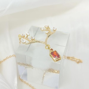 Op de afbeelding: Een goudkleurige ketting met een takontwerp, versierd met heldere kristallen en een rechthoekige, rood-oranje edelsteenhanger. De ketting wordt gepresenteerd op een helder, vierkant blok tegen een witte achtergrond.