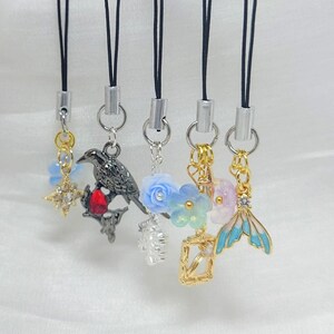 May include: Five phone charms hanging from black cords with silver-tone connectors. Charms include a gold star, a black bird with a red stone, a blue flower, a gold frame with a blue flower, and a blue and gold mermaid tail with a clear stone.