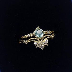 Pode incluir: Um anel dourado com uma pedra quadrada azul clara e pequenas pedras brancas. O anel é apresentado numa caixa azul escura, ideal para oferta.