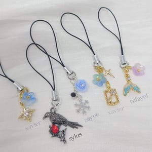 May include: Five phone charms with black cords. Charms include a blue flower with a star, a dark metal raven with a red stone, a blue rose with a snowflake, a blue flower with a cross and frame, and a blue mermaid tail with a purple flower.