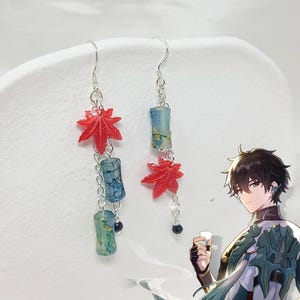 May include: Dangling earrings featuring red maple leaf charms and blue-green cylindrical beads. The earrings are connected by silver chains and hooks. The design includes small black beads at the bottom of the chains. The earrings are displayed against a white background.
