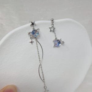 Puede incluir: Un par de pendientes plateados con tem&aacute;tica celestial. Uno presenta un orbe planetario azul y morado, adornado con estrellas y una cadena colgante. El otro tiene un dije de planeta similar y detalles de estrellas. Un accesorio &uacute;nico y elegante.