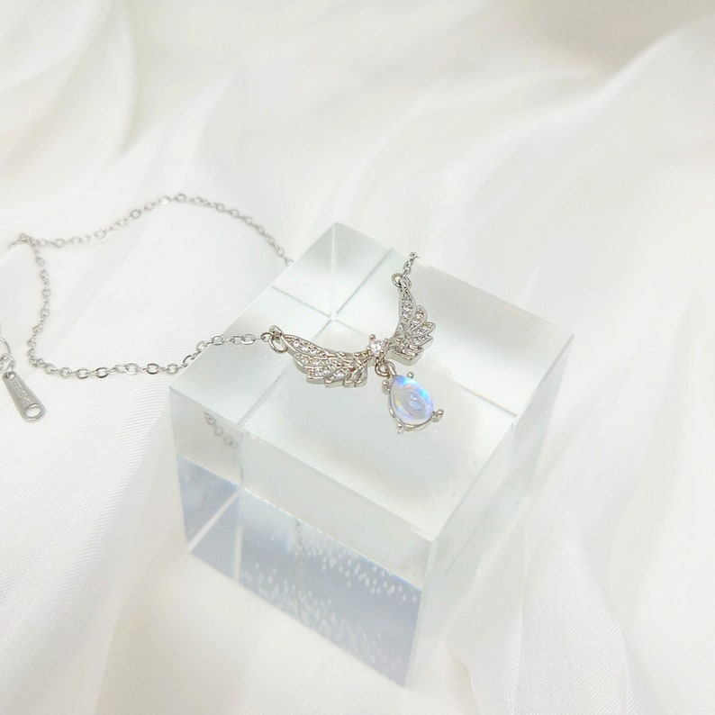 May include: A silver necklace with a teardrop-shaped moonstone pendant and a pair of angel wings adorned with clear cubic zirconia stones.