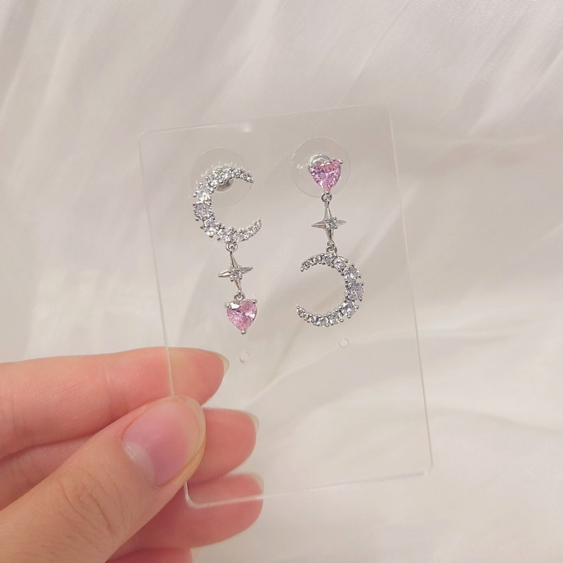 May include: A pair of silver earrings with a crescent moon design, pink heart accents, and clear crystal embellishments.