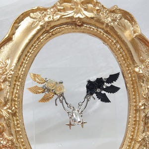 May include: A pair of decorative earrings featuring wing and rose designs. One earring has gold wings and cream roses, the other has black wings and a black rose. The earrings are connected by a silver chain and star charms.