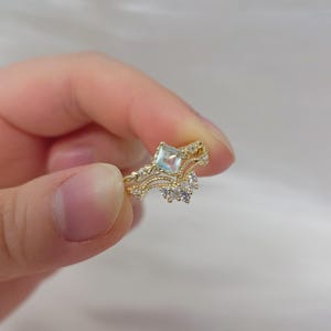 May include: A gold-colored ring with a square, light blue gemstone at the center. The ring features a delicate, ornate design with small, clear accent stones. The ring is held in a person's hand.
