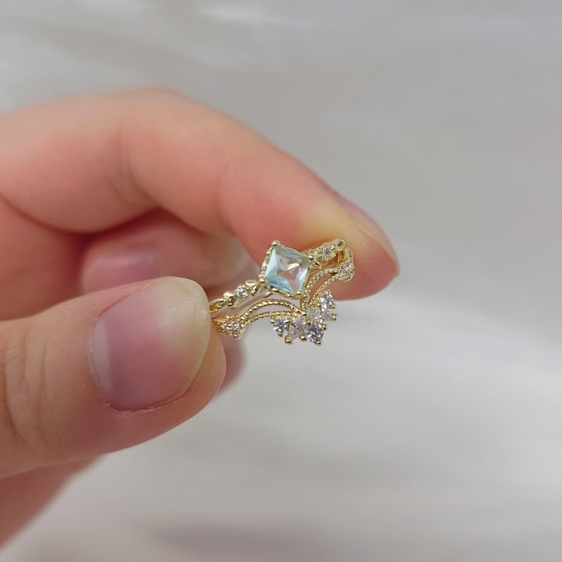 May include: A gold-colored ring with a square, light blue gemstone at the center. The ring features a delicate, ornate design with small, clear accent stones. The ring is held in a person's hand.