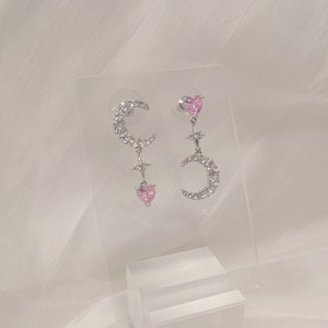 May include: A pair of silver earrings with a crescent moon design, pink heart accents, and clear crystal embellishments.