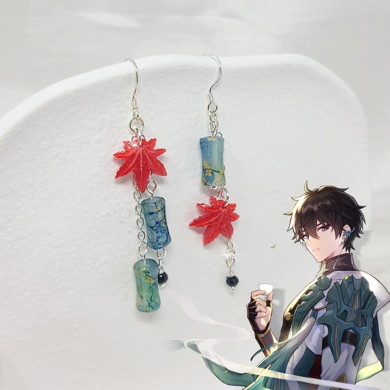 May include: Dangling earrings featuring red maple leaf charms and blue-green cylindrical beads. The earrings are connected by silver chains and hooks. The design includes small black beads at the bottom of the chains. The earrings are displayed against a white background.