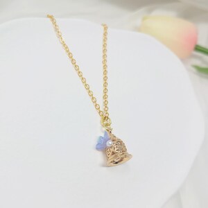 May include: A gold-colored bell pendant necklace with a delicate chain. The bell has intricate cut-out designs, a small pearl, and a light blue flower accent. The necklace is displayed on a white surface.