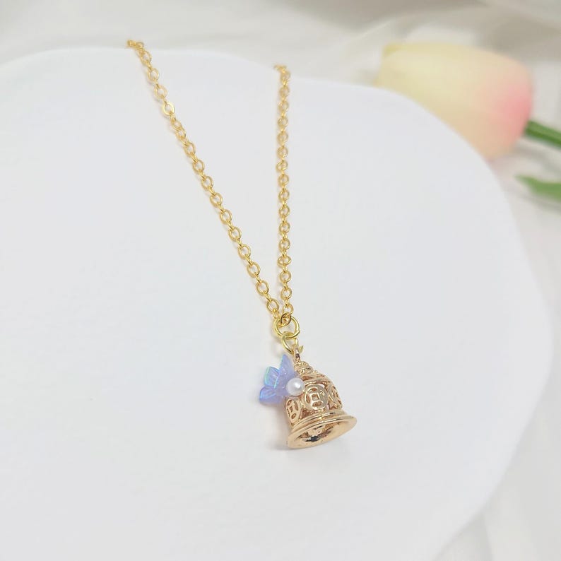 May include: A gold-colored bell pendant necklace with a delicate chain. The bell has intricate cut-out designs, a small pearl, and a light blue flower accent. The necklace is displayed on a white surface.