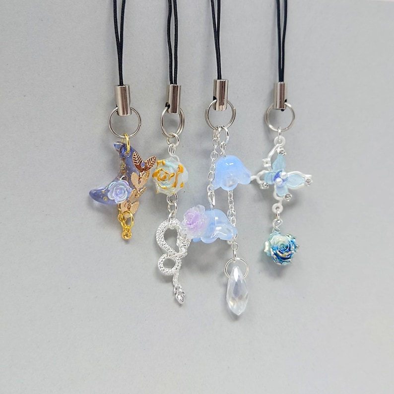 K&ouml;nnte beinhalten: Vier dekorative Handy-Anh&auml;nger mit verschiedenen Designs. Einer zeigt einen blauen Halbmond mit einer Rose, ein anderer eine gelb-wei&szlig;e Blume, ein dritter eine Schlange mit blauen Blumen und einem Kristall, und der letzte einen Schmetterling und eine Rose.