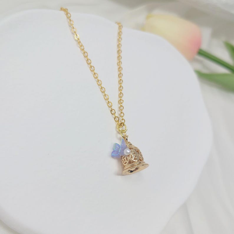 May include: A gold-colored necklace with a bell-shaped pendant. The pendant features a blue butterfly and a small pearl. The necklace chain is also gold-colored. The background is white.