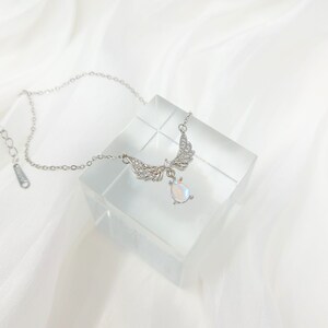 May include: A silver necklace with a teardrop-shaped pendant and angel wings. The pendant is iridescent and has a sparkly finish. The wings are adorned with small clear crystals.