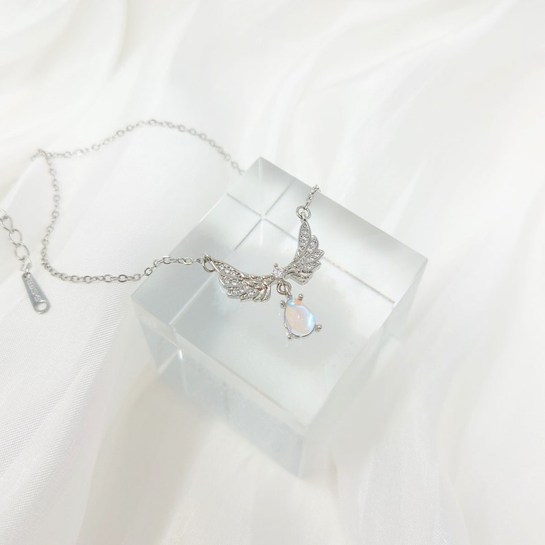 May include: A silver necklace with a teardrop-shaped pendant and angel wings. The pendant is iridescent and has a sparkly finish. The wings are adorned with small clear crystals.