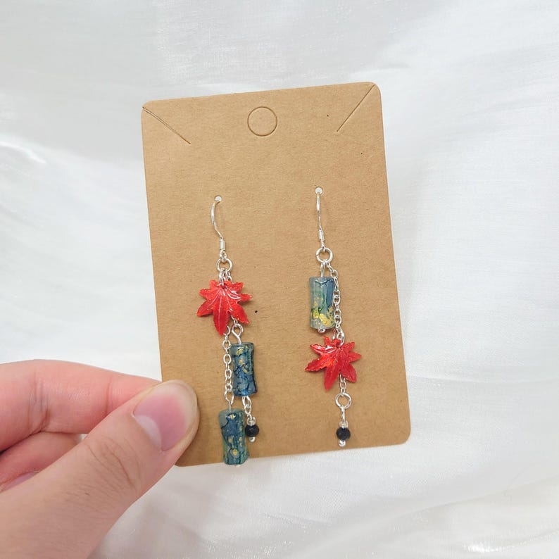 May include: Dangle earrings featuring red maple leaf charms and cylindrical blue and green beads. The earrings are suspended from silver-colored hooks and chains, with a small black bead at the bottom of each earring.