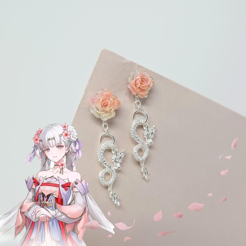 May include: A pair of silver-colored dragon earrings with dangling clear crystals. Each earring features a peach-colored rose at the top. The earrings are displayed on a pink surface with a cartoon character in the background.
