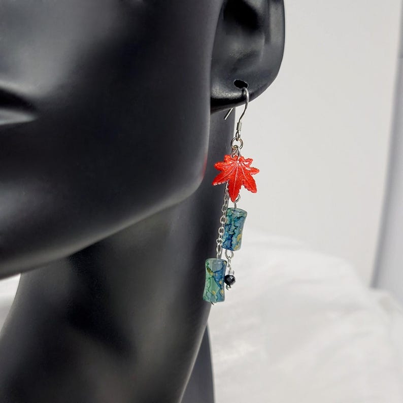 May include: Dangling earrings featuring a red maple leaf charm, blue and green cylindrical beads, and a small black bead. The earrings are suspended from a silver-colored hook. The design is inspired by nature.